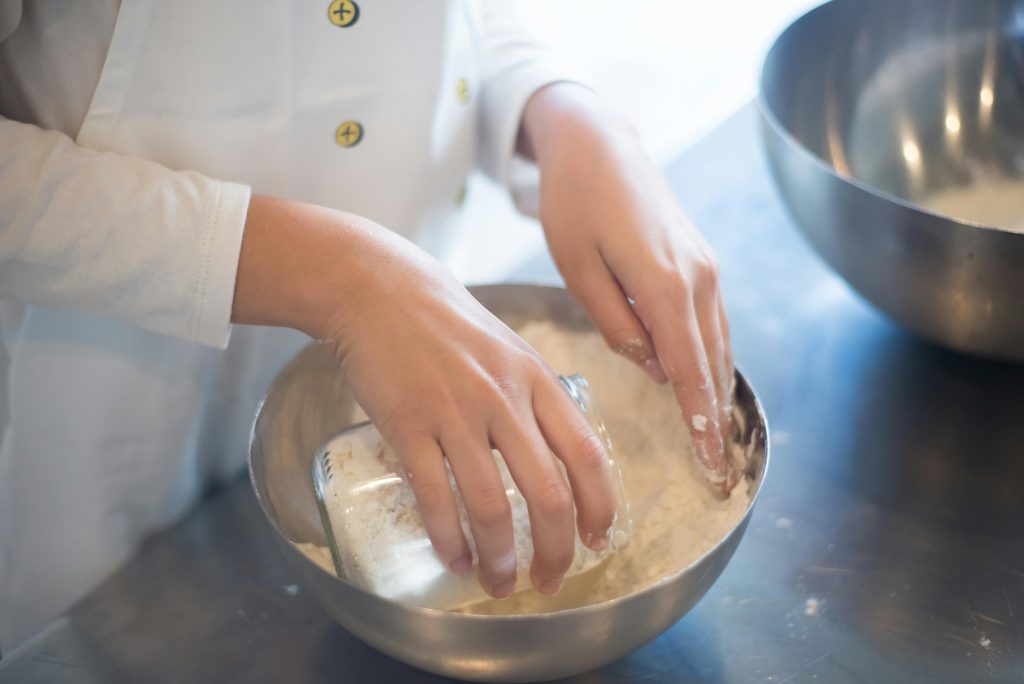 A person wearing a white apron mixes flour in a stainless steel bowl in a kitchen.